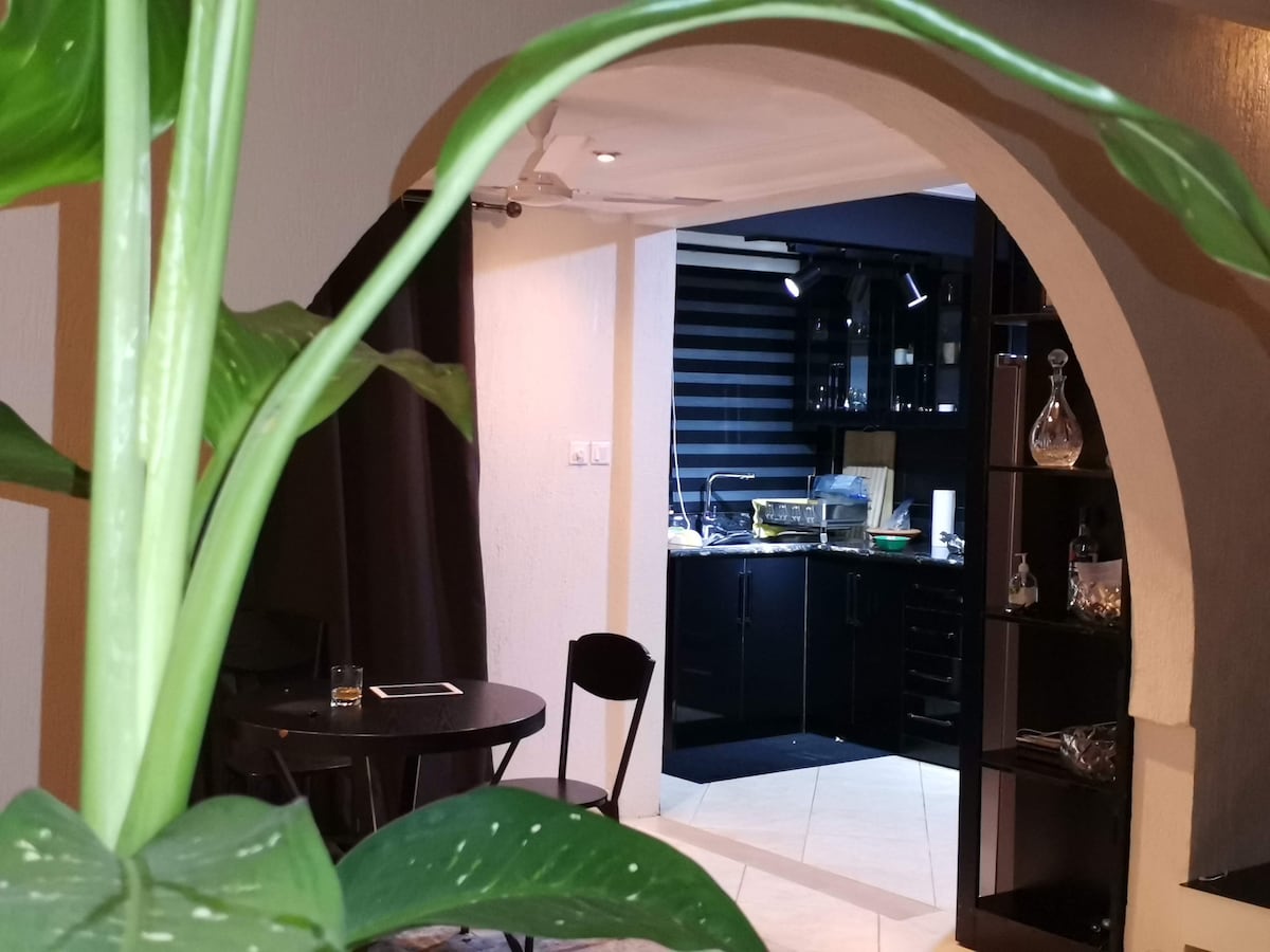 An archway frames a view into the modern kitchen, where sleek black cabinetry and appliances are visible. A small round table with chairs sits in the foreground, accompanied by a lush green plant, adding a touch of natural color to the scene.