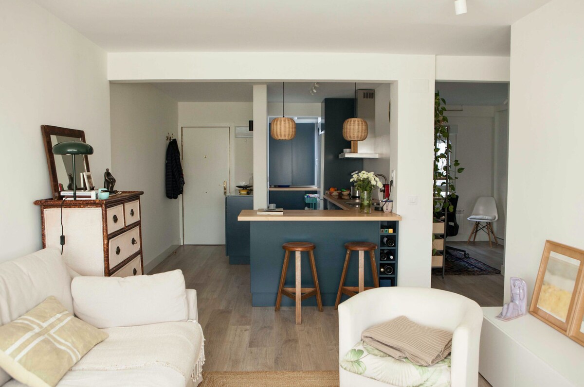 A bright, open-plan living space features a modern kitchen island with bar stools. Soft furnishings include a light sofa and an inviting throw. Wooden elements are complemented by natural light streaming through large windows, enhancing the room's spacious feel.