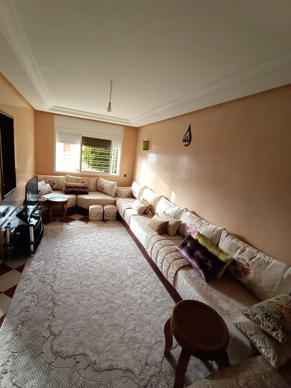 A light-filled living room features a spacious sofa adorned with various cushions, arranged along two walls. A decorative rug covers the floor, and a small wooden table is positioned nearby. Natural light filters through a window, highlighting the warm tones of the walls.