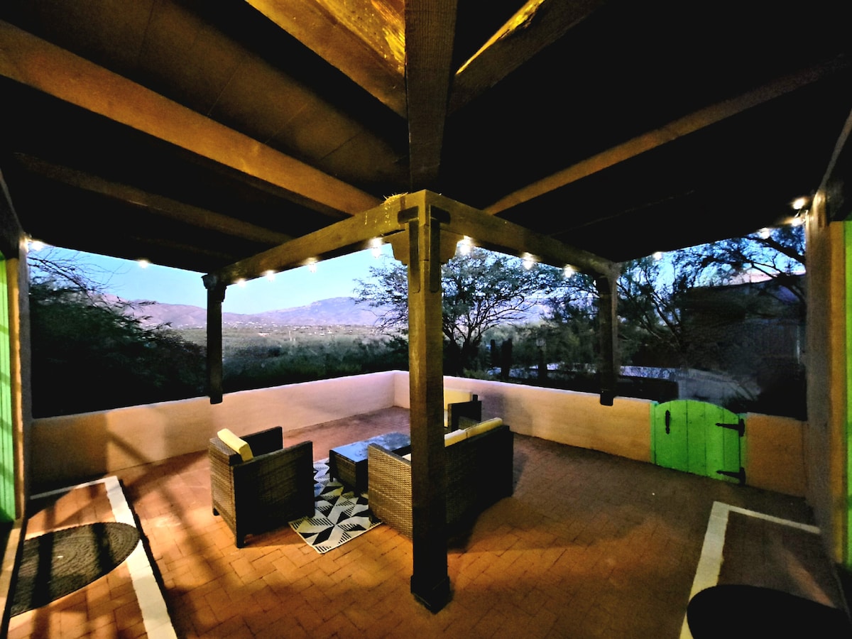 A covered outdoor patio area is presented, featuring comfortable seating arrangements around a low table. Soft lighting enhances the space while abundant views of the surrounding mountains and trees are visible in the background.