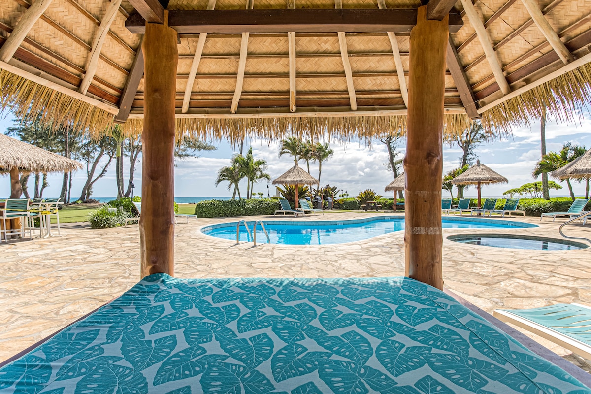A thatched-roof cabana provides shade overlooking a resort pool and palm trees. The pool, surrounded by decorative stone work, is visible along with several Bali style cabanas. Guests can enjoy the serene ocean views beyond the pool area.