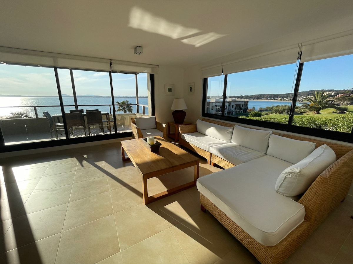 A spacious living area features large windows offering unobstructed views of the ocean and surrounding landscape. A comfortable light-colored sectional sofa is positioned around a wooden coffee table. Natural light floods the space, highlighting the serene ambiance.