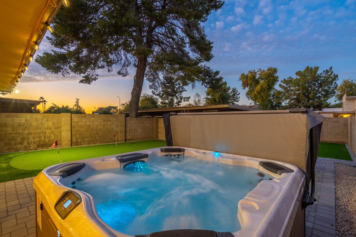 A spacious outdoor jacuzzi is filled with bubbling water, illuminated by soft blue lights. The surrounding area features a well-maintained lawn, a putting green, and a large tree, with the evening sky showcasing a gradient of soft hues.