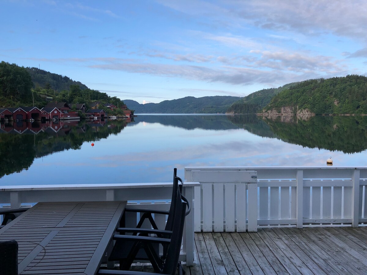 A peaceful view is presented from the waterfront deck, featuring a wooden table and chairs. The calm water reflects the surrounding hills and clouds, with charming red cabins visible along the shoreline in the distance.