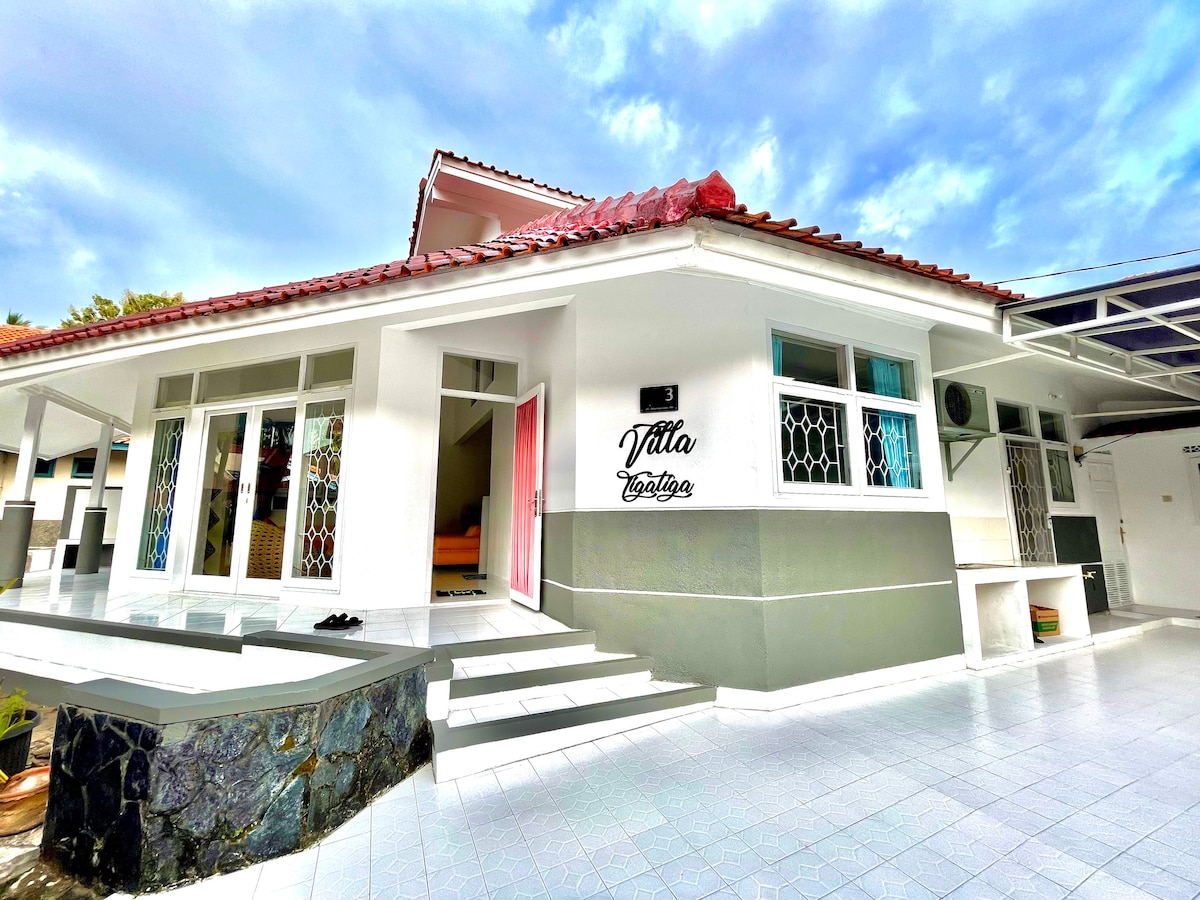The exterior of Villa Tigatiga is presented, showcasing a modern architectural design with white walls and a red-tiled roof. Large glass doors provide natural light, while a spacious patio area is visible, surrounded by a well-maintained surface and greenery in the background.
