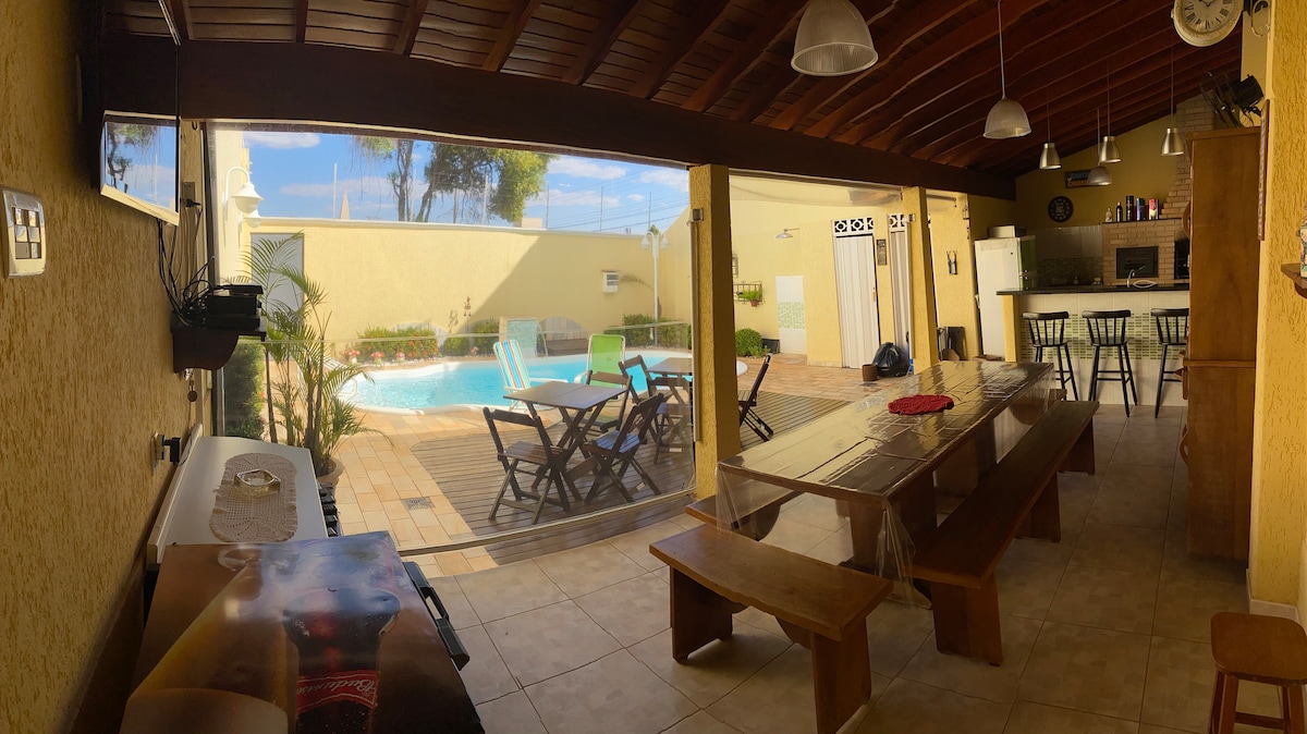 A spacious indoor area offers a view of the outdoor pool through large sliding glass doors. Comfortable seating is arranged at a large wooden table with additional chairs nearby. Bright lighting and a rustic wooden ceiling enhance the airy atmosphere.