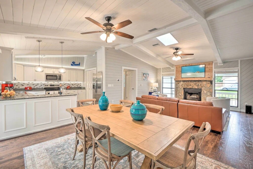 An open living space features a wooden dining table with light blue vases, accented by a cozy seating area with a tan sofa. The kitchen is visible in the background, equipped with modern appliances and a stone fireplace nearby. Natural light is captured through the skylight.