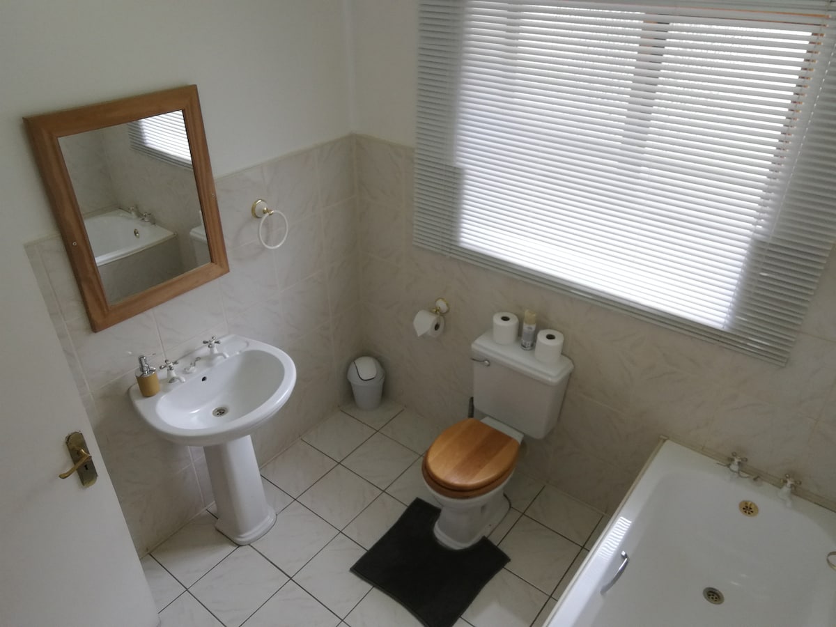 A well-appointed bathroom is displayed, featuring a sink with a wooden-framed mirror above. A toilet with a wooden seat, alongside a bathtub, is positioned in the space. Window blinds provide privacy while allowing natural light to enter.