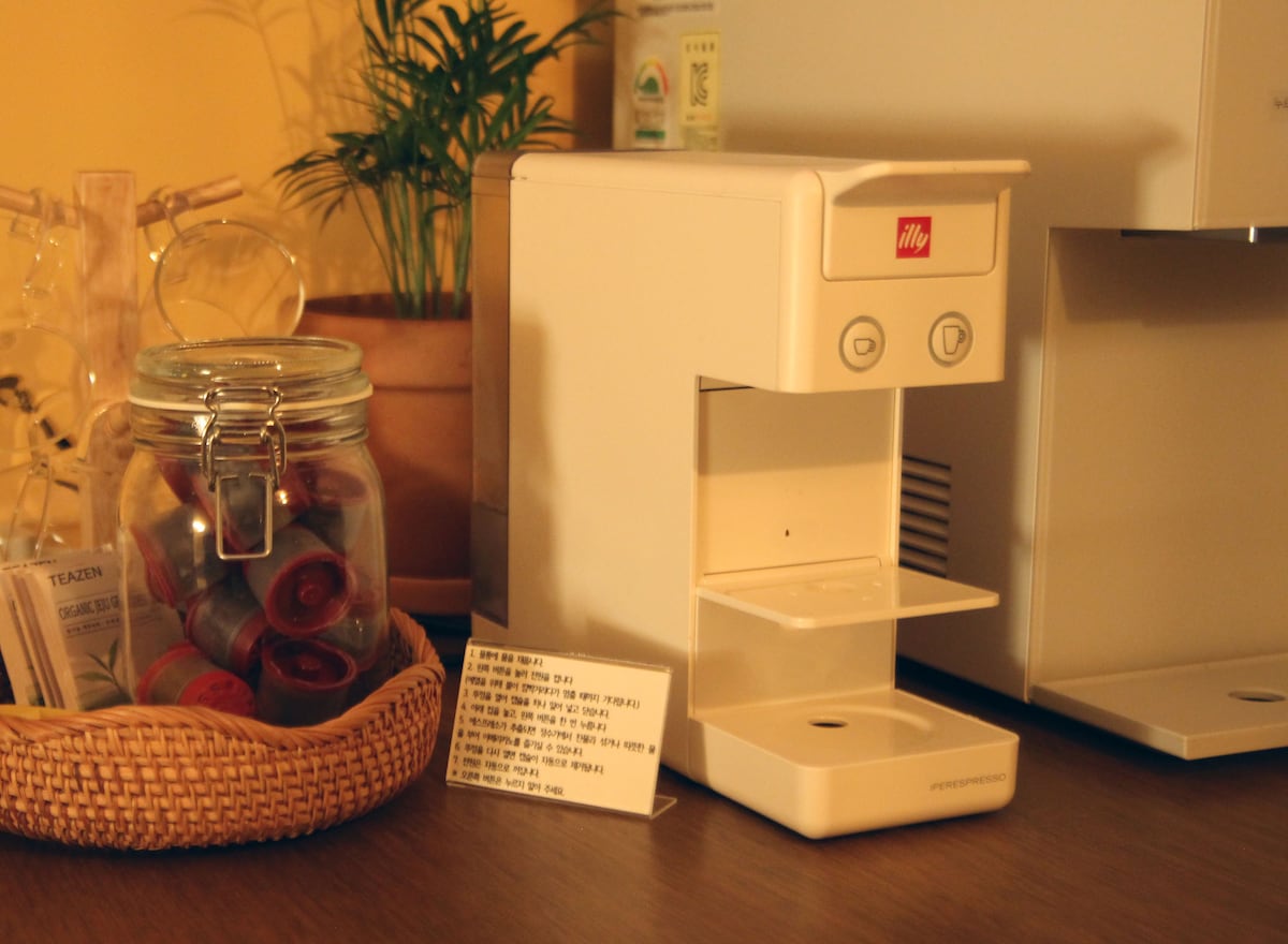 An illy capsule coffee machine is placed on a wooden surface, next to a glass jar filled with colorful coffee capsules. A small woven basket contains various tea bags, alongside a card detailing the offerings.