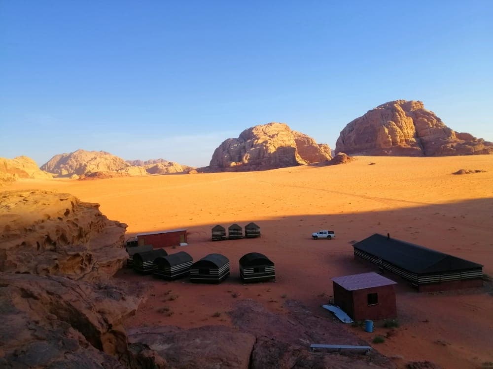 A desert landscape features a cluster of traditional Bedouin tents nestled in a sandy area. Rocky mountains rise in the backdrop under a clear blue sky, casting a gentle shadow across the ground. A vehicle is positioned nearby, emphasizing the remote tranquility of the location.