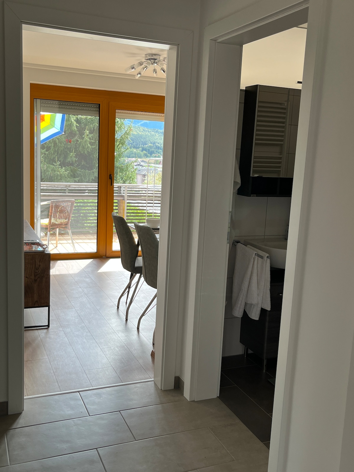 A bright hallway leads to an open space featuring a dining area with natural light pouring in through large glass doors. Outdoor seating is visible on the balcony, showcasing views of the surrounding landscape.