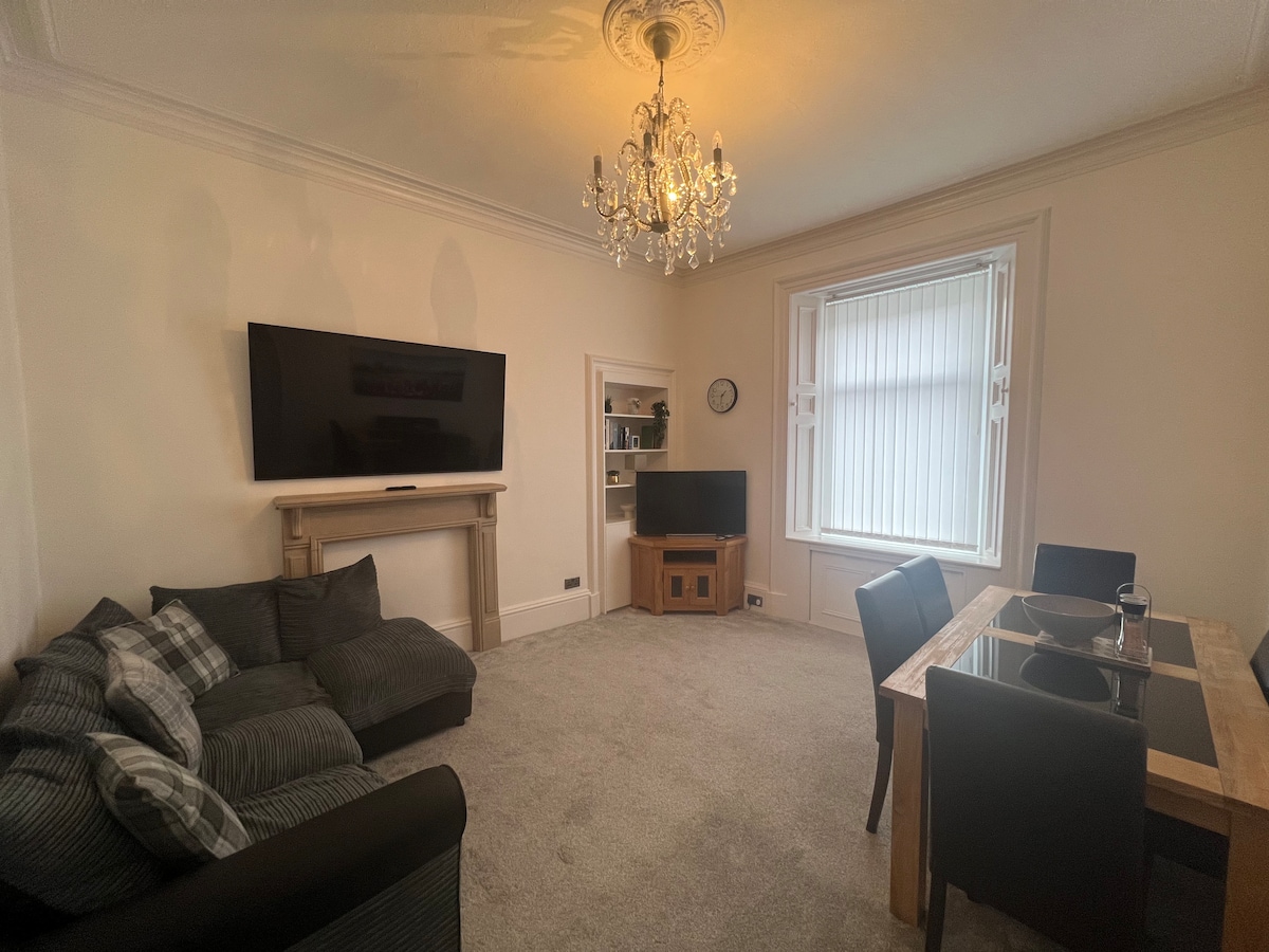 A spacious living area features a large sectional sofa with patterned cushions and a wooden dining table accompanied by black chairs. Two television sets are positioned in the room, and natural light filters through a large window with vertical blinds.