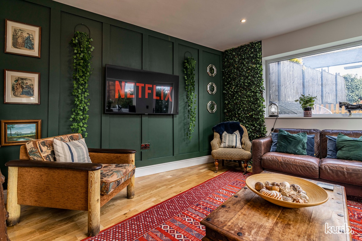 A cozy living room features a leather sofa and an armchair set against a green accent wall adorned with plants. A large, wall-mounted Smart TV displays the Netflix logo, while a decorative coffee table holds a bowl of shells. Natural light streams in through a window, illuminating a patterned rug.