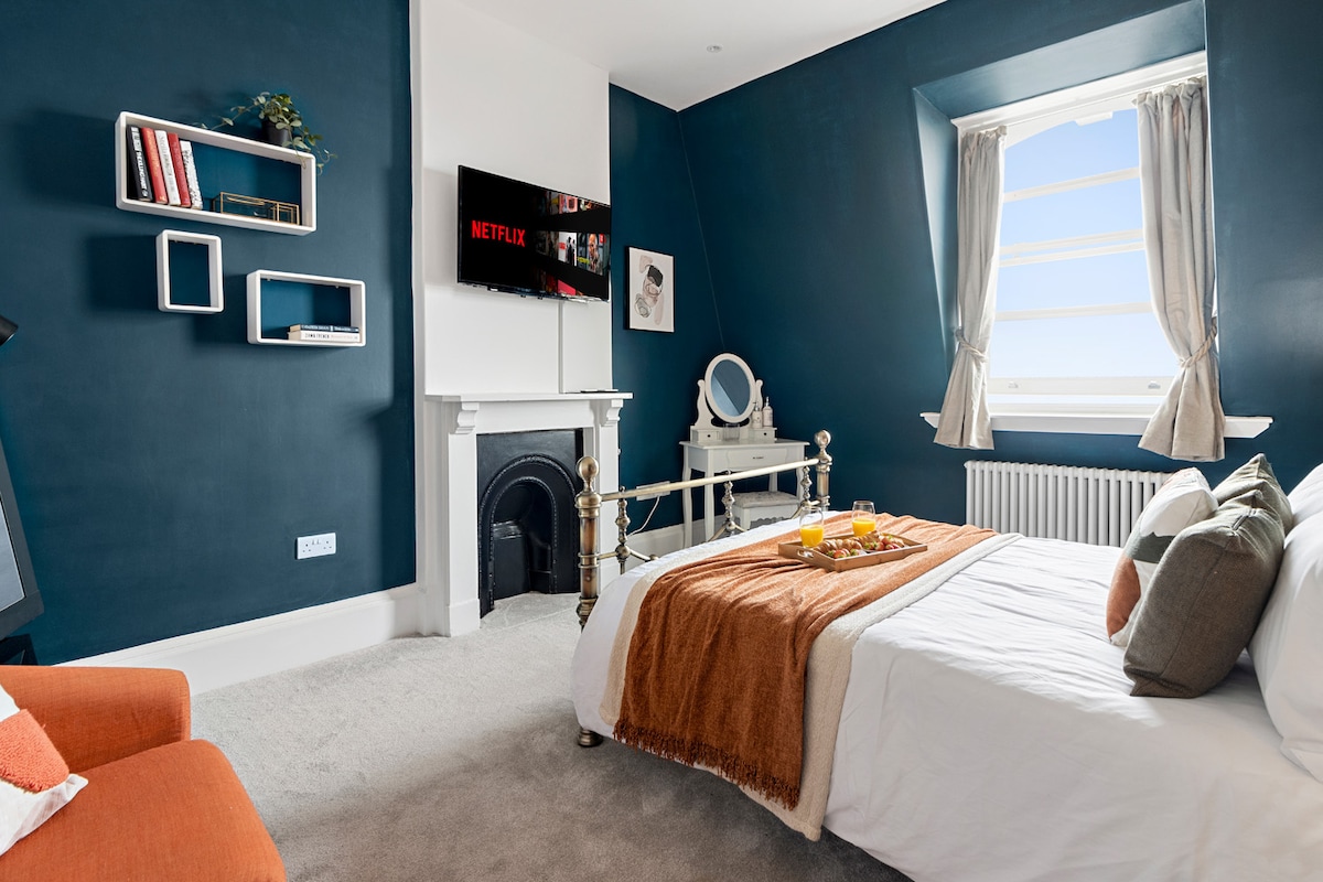 The vibrant sleeping area features a spacious bed adorned with a soft throw blanket and decorative pillows. A wall-mounted TV provides access to Netflix. A cozy armchair complements the room, alongside a small dresser and a mirror. A fireplace adds character to the space.
