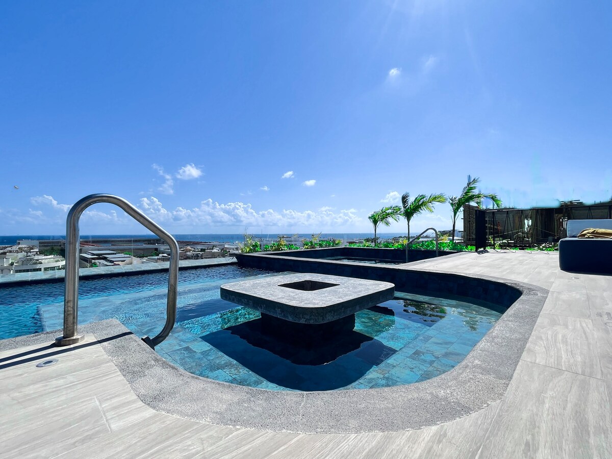 A rooftop pool area is displayed, featuring a square spa surrounded by a spacious pool. Lush greenery accents the landscape, while a clear blue sky and distant ocean views create a serene backdrop.