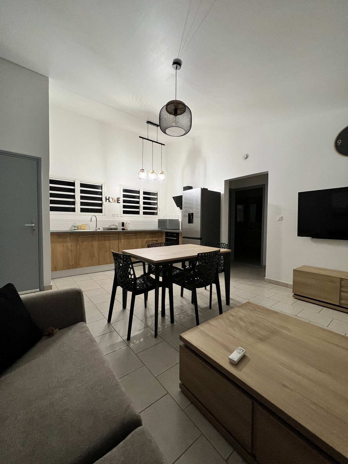 An open living space is presented, featuring a dining table surrounded by four chairs, positioned near a modern kitchen. The kitchen includes sleek cabinetry and appliances, and a television is mounted on the wall opposite the seating area, contributing to a functional layout.