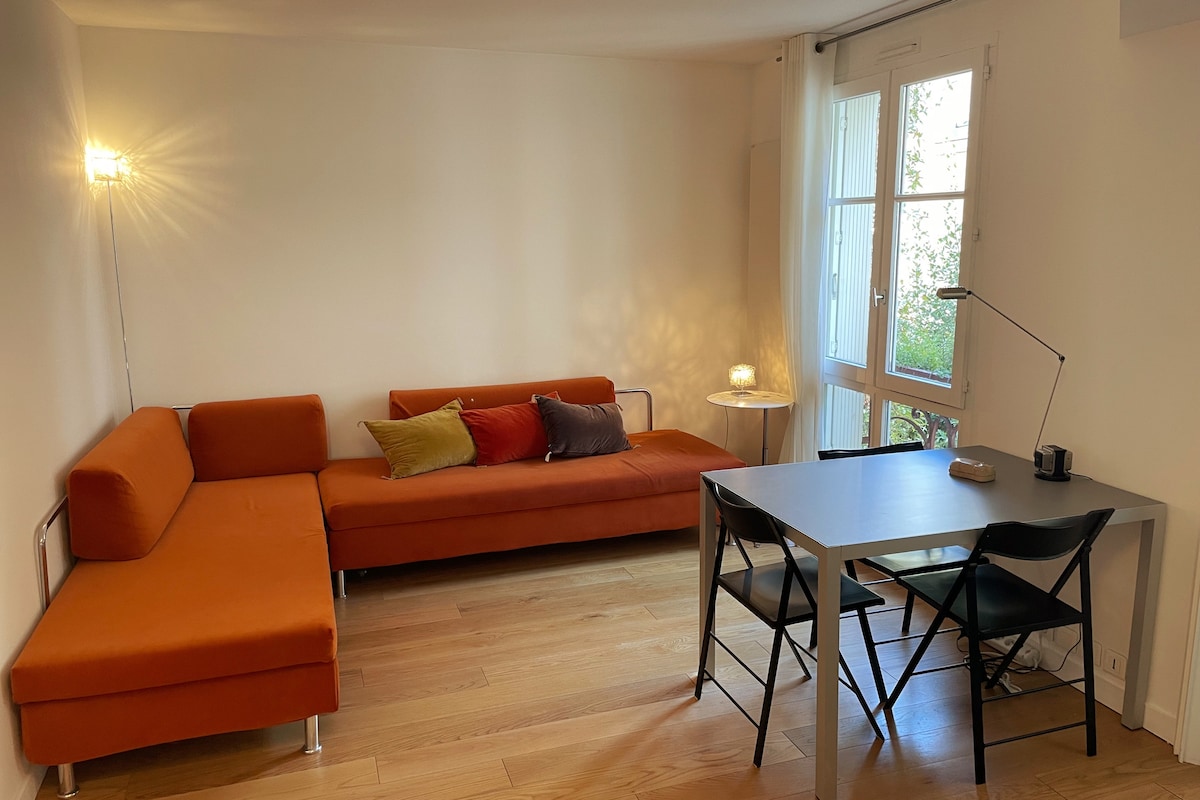 The living area features two orange sofas arranged for comfort, with decorative pillows adding color. A small side table is present, alongside a stylish desk with four black chairs. Natural light filters through large windows, enhancing the space's brightness.