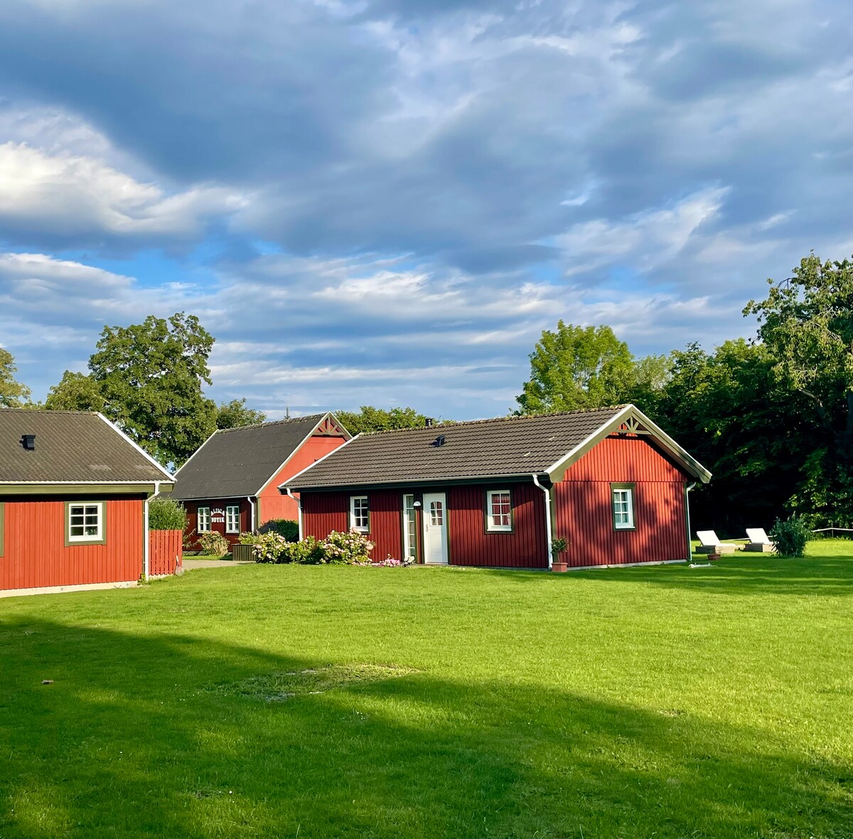 A cluster of charming red wooden houses is set against a backdrop of green grass and trees. The structures feature distinct roofs and white-framed windows, creating a welcoming appearance. A landscaped garden area enhances the natural surroundings.