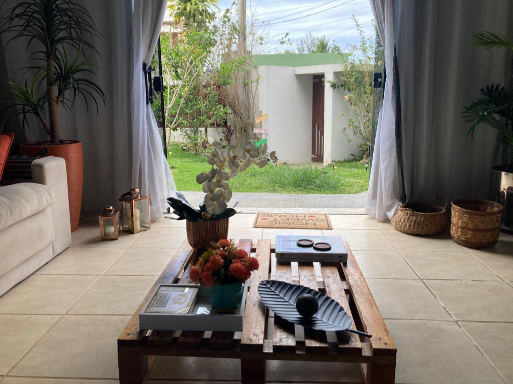 A cozy interior view is presented, featuring a wooden coffee table adorned with plants and decorative items. Natural light filters through sheer curtains, illuminating the space. A view of the greenery outside is visible, enhancing the connection to nature.