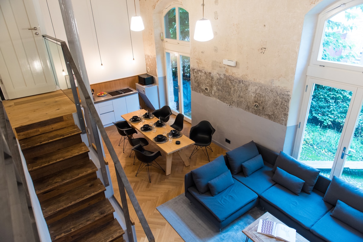 An aerial view of the open living area showcases a spacious seating arrangement with a large gray sectional sofa. Nearby, a wooden table is set for dining, surrounded by black chairs. Natural light floods the area through multiple large windows, enhancing the modern kitchen visible in the background.