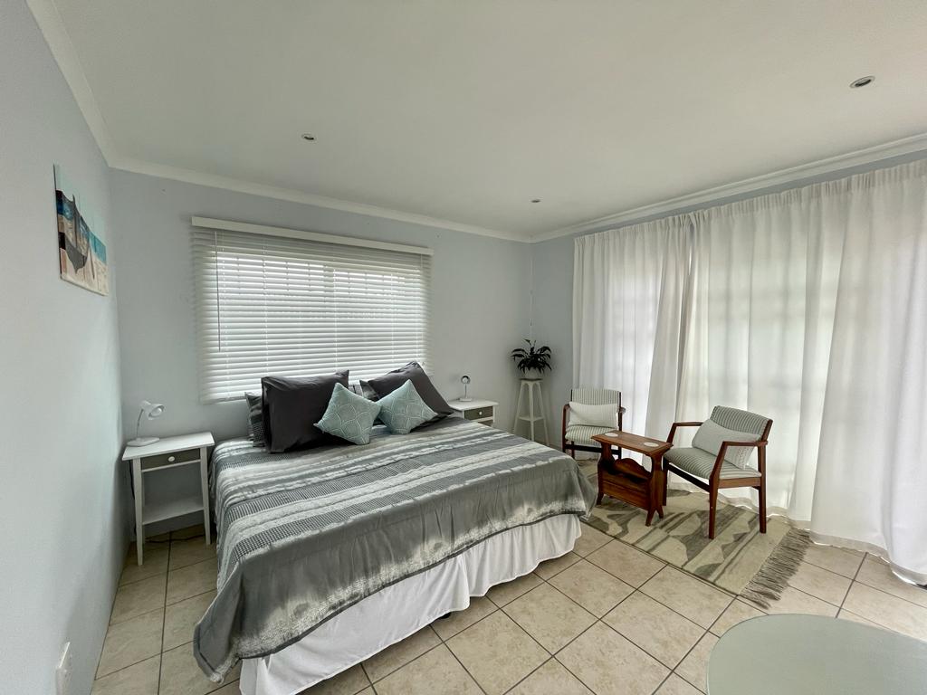 An open plan bedroom and living area features a spacious bed adorned with neutral bedding and decorative pillows. Natural light filters through a wide window, complemented by sheer curtains. A cozy seating arrangement includes two chairs and a wooden table, offering a relaxed ambiance.