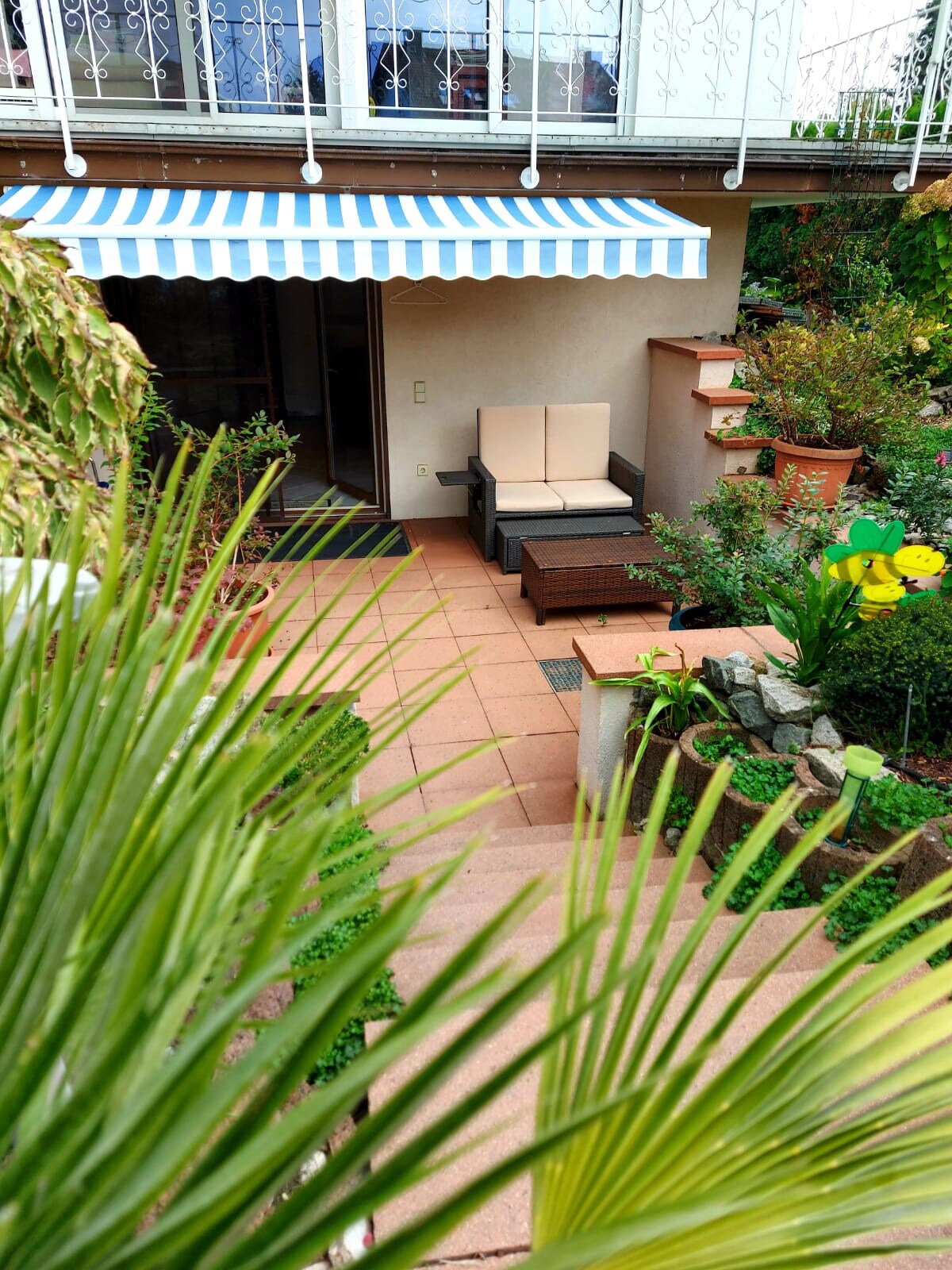 A charming terrace is adorned with potted plants and surrounding greenery. A striped awning offers shade over two comfortable chairs, positioned next to a small table. The area invites relaxation and enjoyment of the outdoors, showcasing a well-maintained garden space.