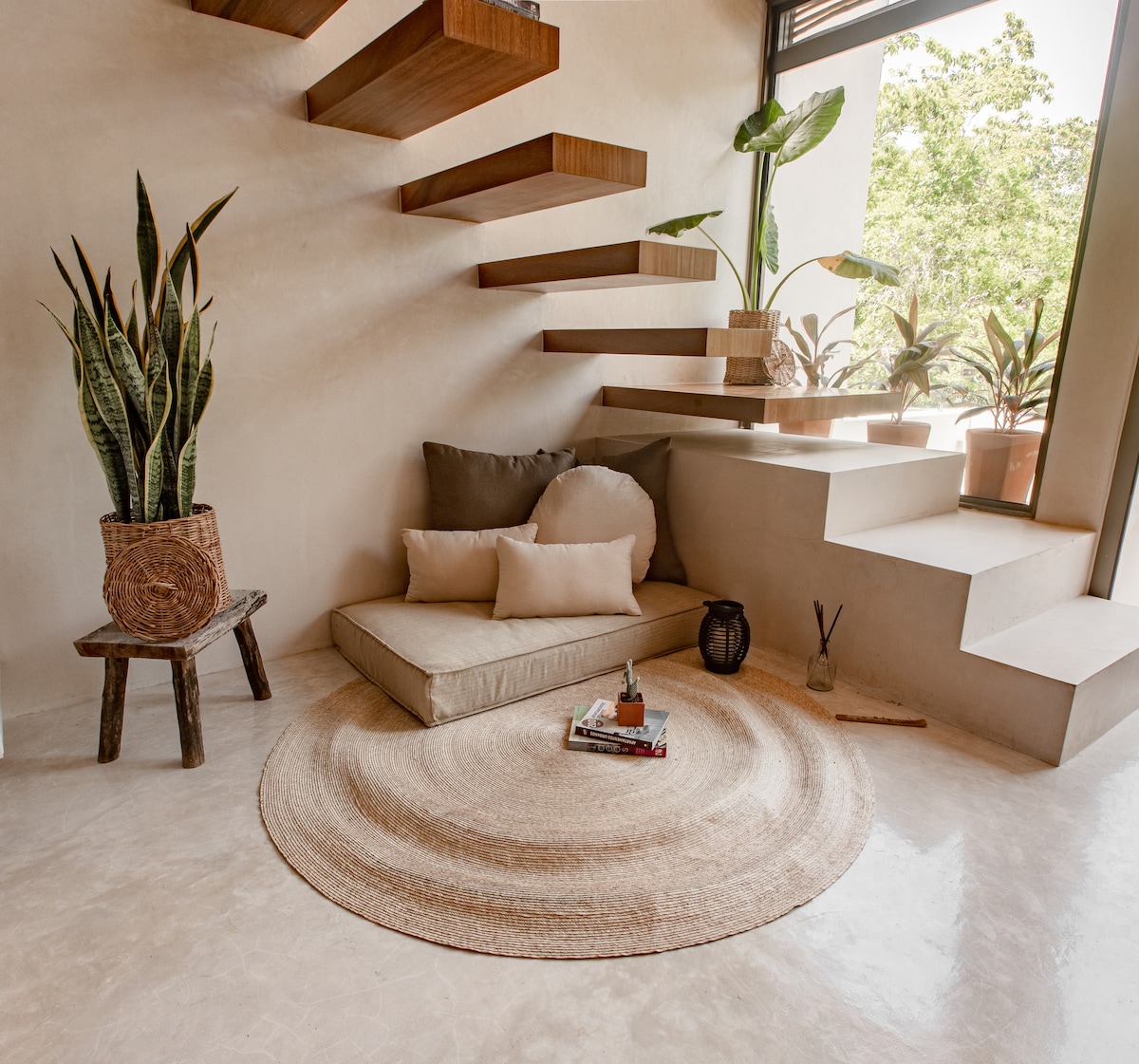A serene indoor corner features a custom-made lounging area with plush pillows and a tasteful round rug. Decorative plants are positioned around the space, while warm wooden stairs ascend gracefully. Natural light filters through large windows, illuminating the neutral-toned surroundings.