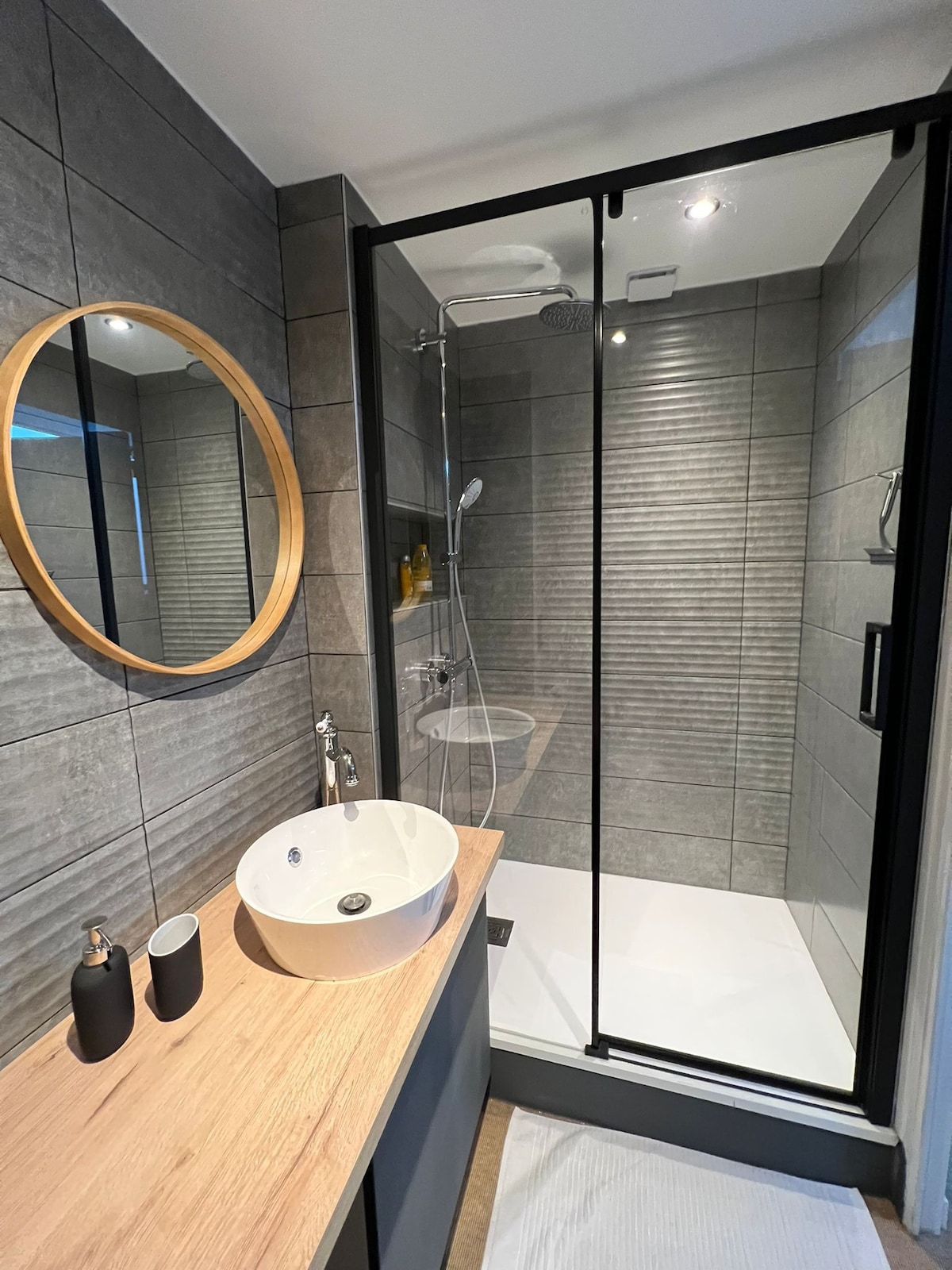 A modern bathroom features a glass shower enclosure with a textured wall design. A round mirror above a wooden vanity complements a stylish sink. A minimalistic arrangement of black and white accessories is displayed on the counter, enhancing the sleek and clean aesthetic.