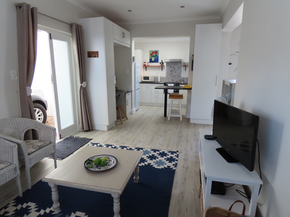 An open-plan lounge area features a light wood floor and a large window allowing natural light. A comfortable couch complements a coffee table, while a television is positioned on a white cabinet. The kitchen is visible in the background with modern appliances.