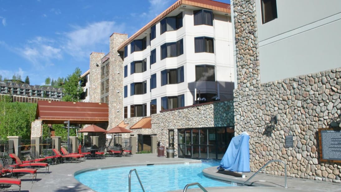 An outdoor pool is visible, surrounded by stone walls and lined with red lounge chairs. Sun umbrellas provide shade, and a small spa area is present. The building's exterior showcases a mix of stone and large windows, reflecting a clear blue sky.