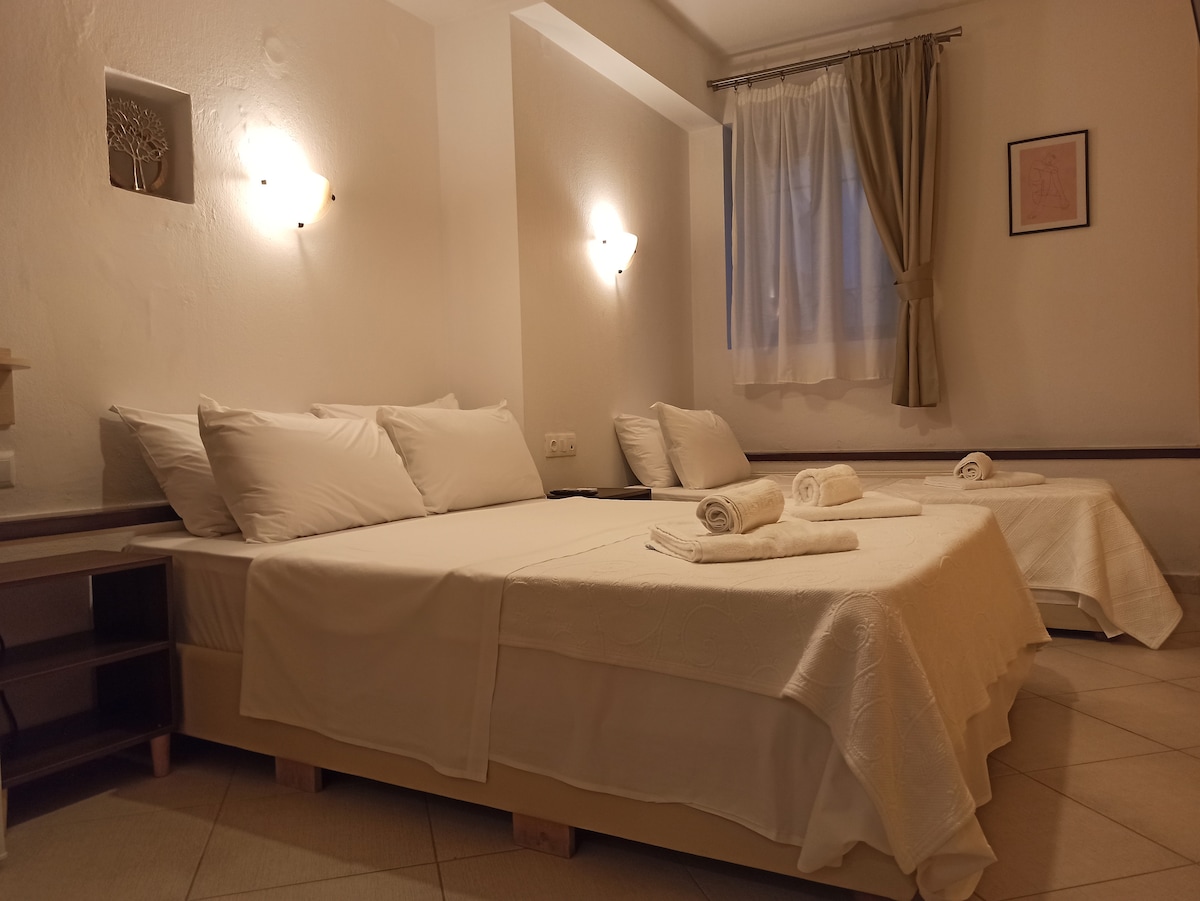 A serene sleeping area features a spacious bed with crisp white linens. Two additional beds are positioned nearby, neatly made with white blankets. Soft lighting is provided by wall-mounted sconces, and natural light enters through a window dressed with sheer curtains.