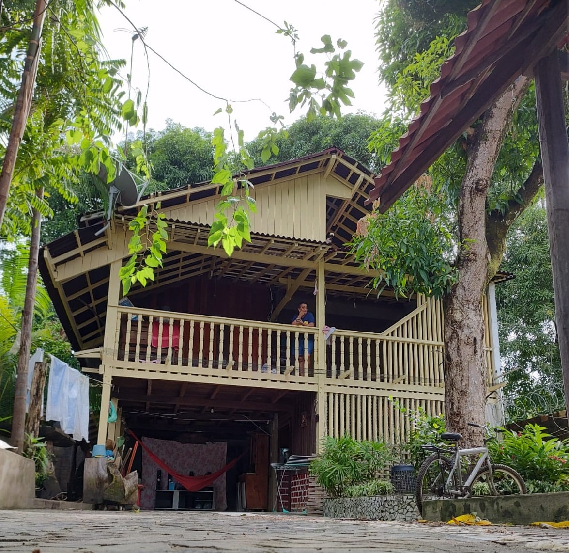 A charming chalet-style house sits elevated on stilts, surrounded by lush greenery. A spacious balcony is visible, with wooden railings inviting relaxation. Adjacent trees provide shade, enhancing the natural setting. A bicycle rests nearby, adding to the outdoor ambiance.