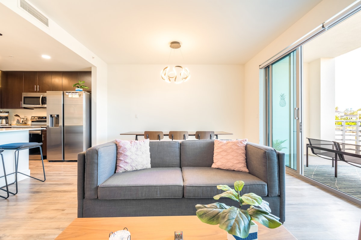 A spacious living area features a modern couch with light pink cushions, facing a dining table that seats six. Large glass doors open to a balcony view, allowing natural light to fill the room. A decorative plant sits on the coffee table.