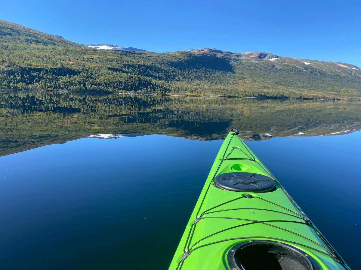 A vibrant green kayak is positioned in calm waters, reflecting the surrounding landscape. The serene lake is bordered by lush green hills and distant mountains, with a clear blue sky overhead. The image captures the tranquility of nature, ideal for outdoor enthusiasts.