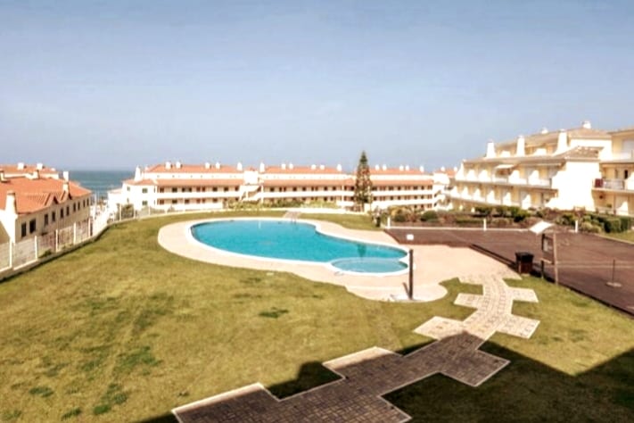 A spacious outdoor area features a large pool with a unique shape, surrounded by manicured green grass. Nearby, well-maintained buildings with red-tiled roofs are visible against a clear sky, providing a serene view of the ocean in the distance.