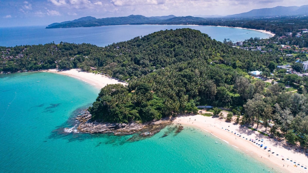 An aerial view captures the stunning coastline, featuring turquoise waters gently lapping against soft sandy beaches. Lush greenery lines the shore, with beachgoers dotted along the shoreline and beach chairs arranged under umbrellas, providing a serene atmosphere for relaxation.