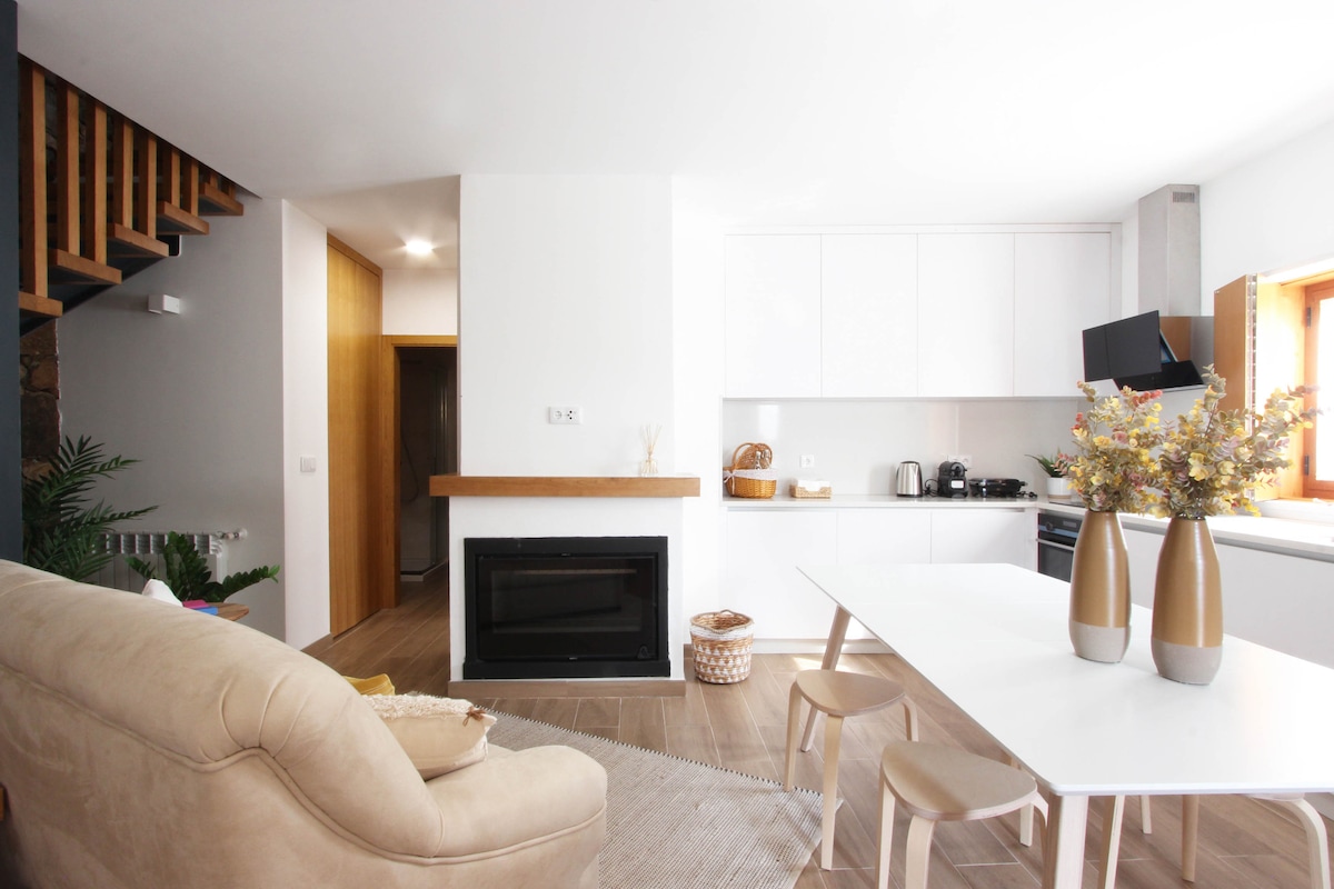 A modern living space features a light-colored sofa paired with a white dining table and wooden chairs. A fireplace is positioned beneath a wooden shelf. The kitchen area includes sleek cabinets and a window that illuminates the room, with decorative vases adding a touch of elegance.