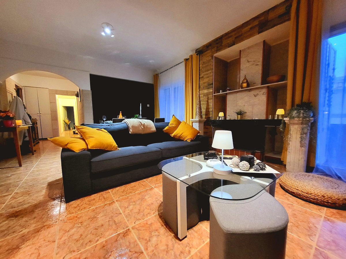A bright living area features a dark sofa adorned with yellow throw pillows. A contemporary coffee table is centrally placed, complemented by decorative items. Soft lighting emanates from a lamp, and large windows allow natural light to enter, enhancing the warm ambiance of the space.