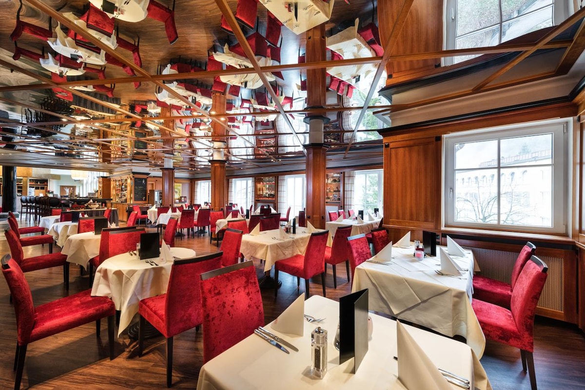 The restaurant features elegant wooden paneling and a reflective ceiling. Tables are set with white linens, accompanied by plush red chairs. Natural light streams through large windows, enhancing the welcoming atmosphere. Each table is equipped with menus and condiments, ready for dining guests.