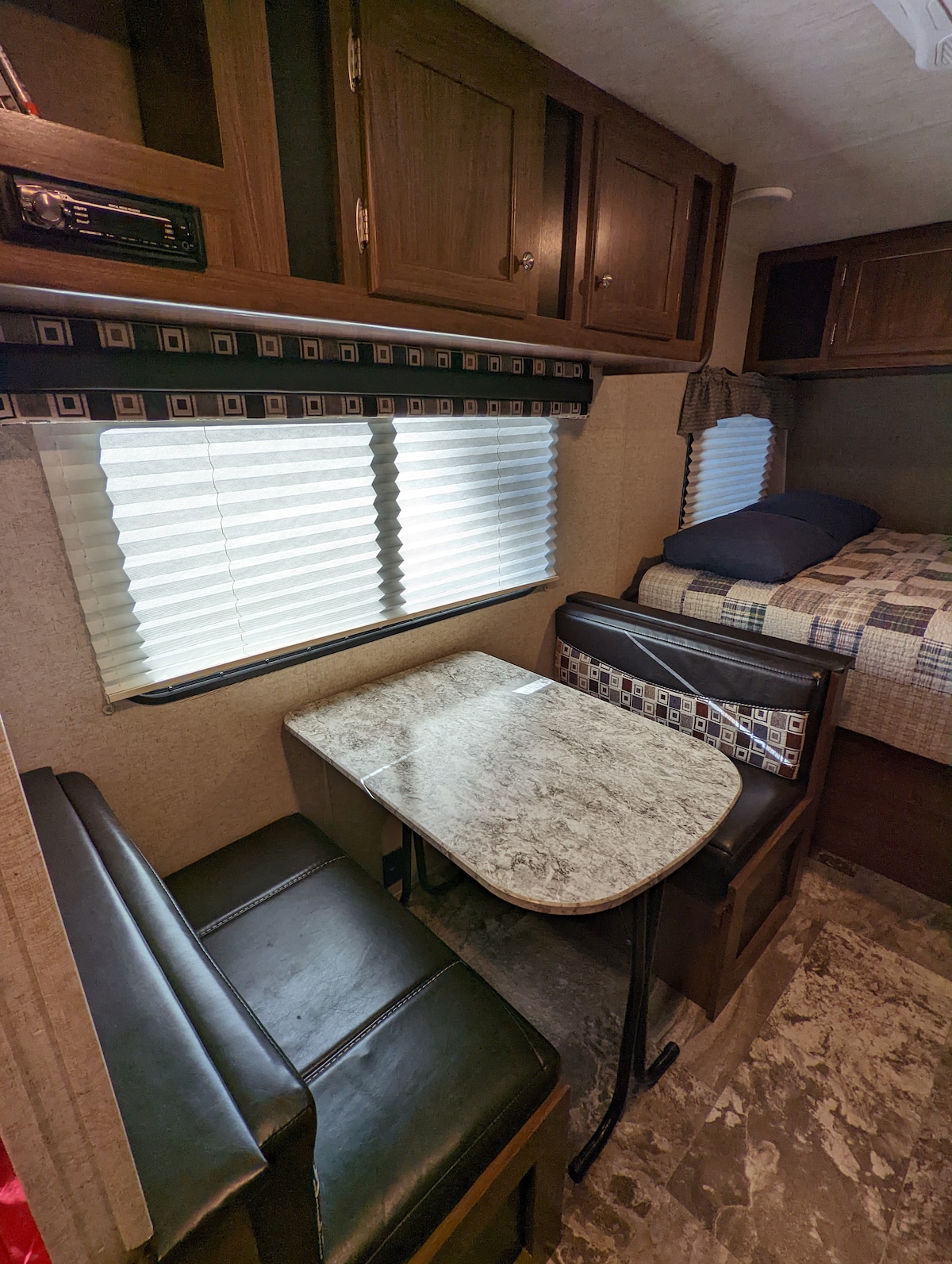 A cozy dinette area features a small table with two cushioned seats, designed for comfort. Natural light filters through the closed blinds, illuminating the space. In the background, a neatly made sleeping area is visible, with a plaid-patterned bedding on a spacious bed.