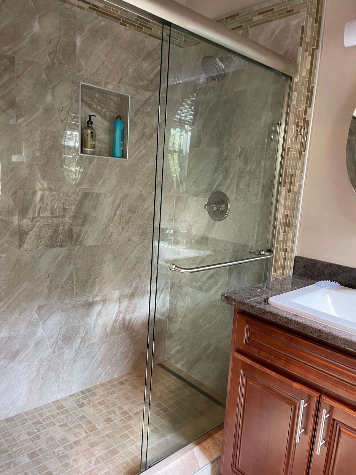 A spacious shower area is enclosed with clear glass, featuring natural stone tile flooring. A built-in shelf holds shower essentials, while modern fixtures are positioned on a smooth tile wall. A sink with a dark countertop and wooden cabinetry is visible adjacent to the shower.