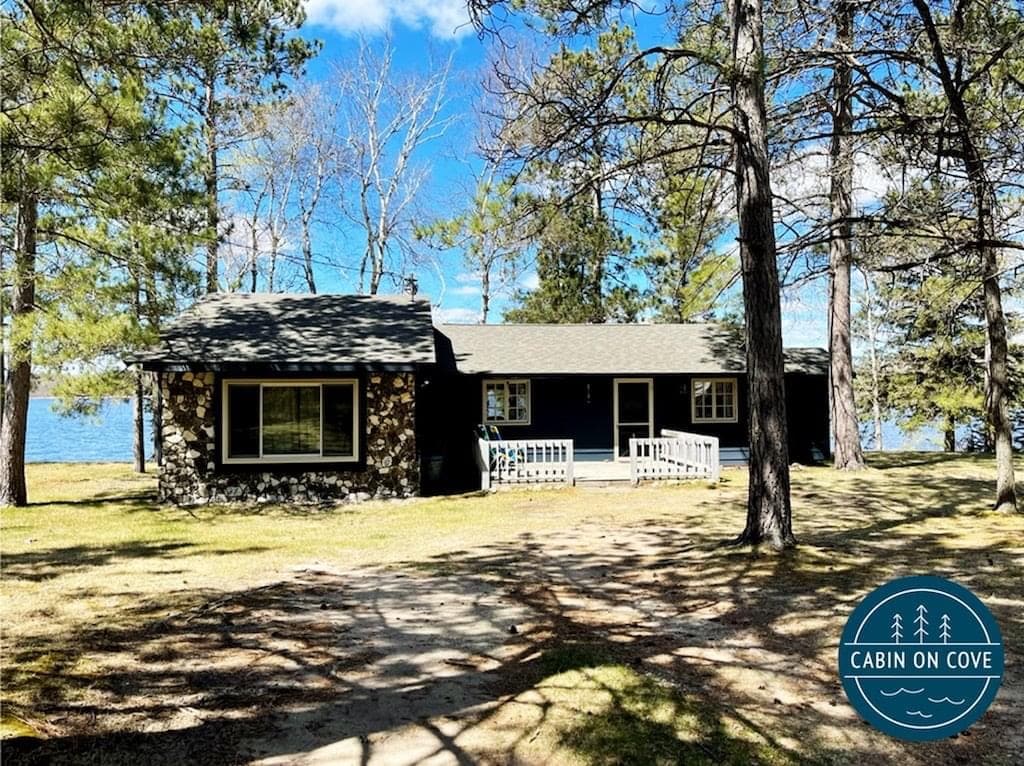 A charming cabin sits under a partly cloudy sky, surrounded by tall trees and a well-maintained lawn. The structure features a mix of stone and wood, with a welcoming porch area and large windows offering views of the nearby lake.