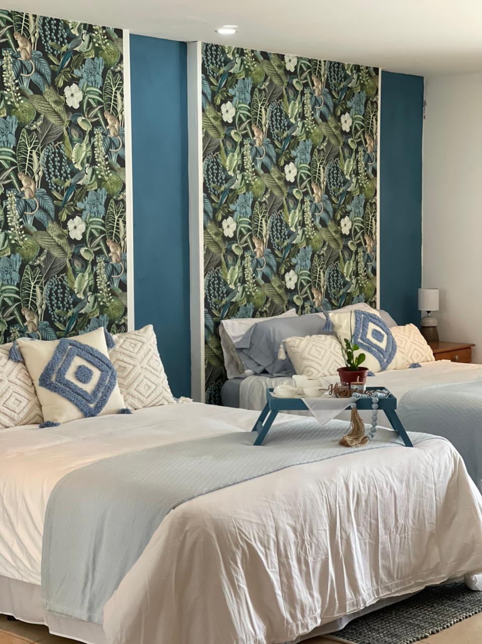 A bright sleeping area features two spacious beds dressed in soft white linens. Behind them, a vibrant botanical wallpaper creates a lively backdrop. A small nightstand holds a lamp, and a tray with a plant is placed on the beds, adding a touch of elegance.
