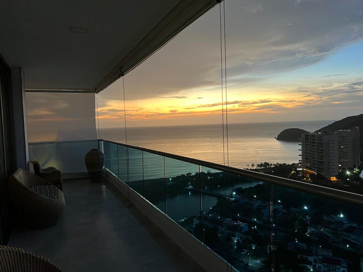 A balcony offers a sweeping view of the ocean and sunset, with the sky illuminated in warm hues. Two woven seating areas are positioned along the edge, providing a comfortable space to enjoy the serene landscapes. The glass railing enhances the panoramic vista.