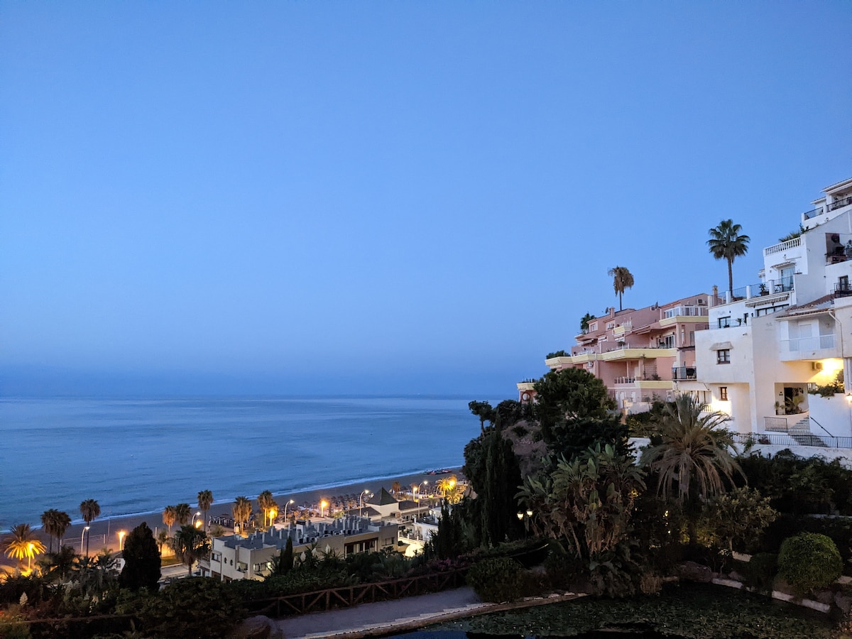 A serene twilight view captures the Mediterranean Sea stretching into the horizon, with soft blue hues above. The coastal town of Nerja is visible, featuring twinkling lights from buildings and palm trees, set against the backdrop of gently undulating cliffs.