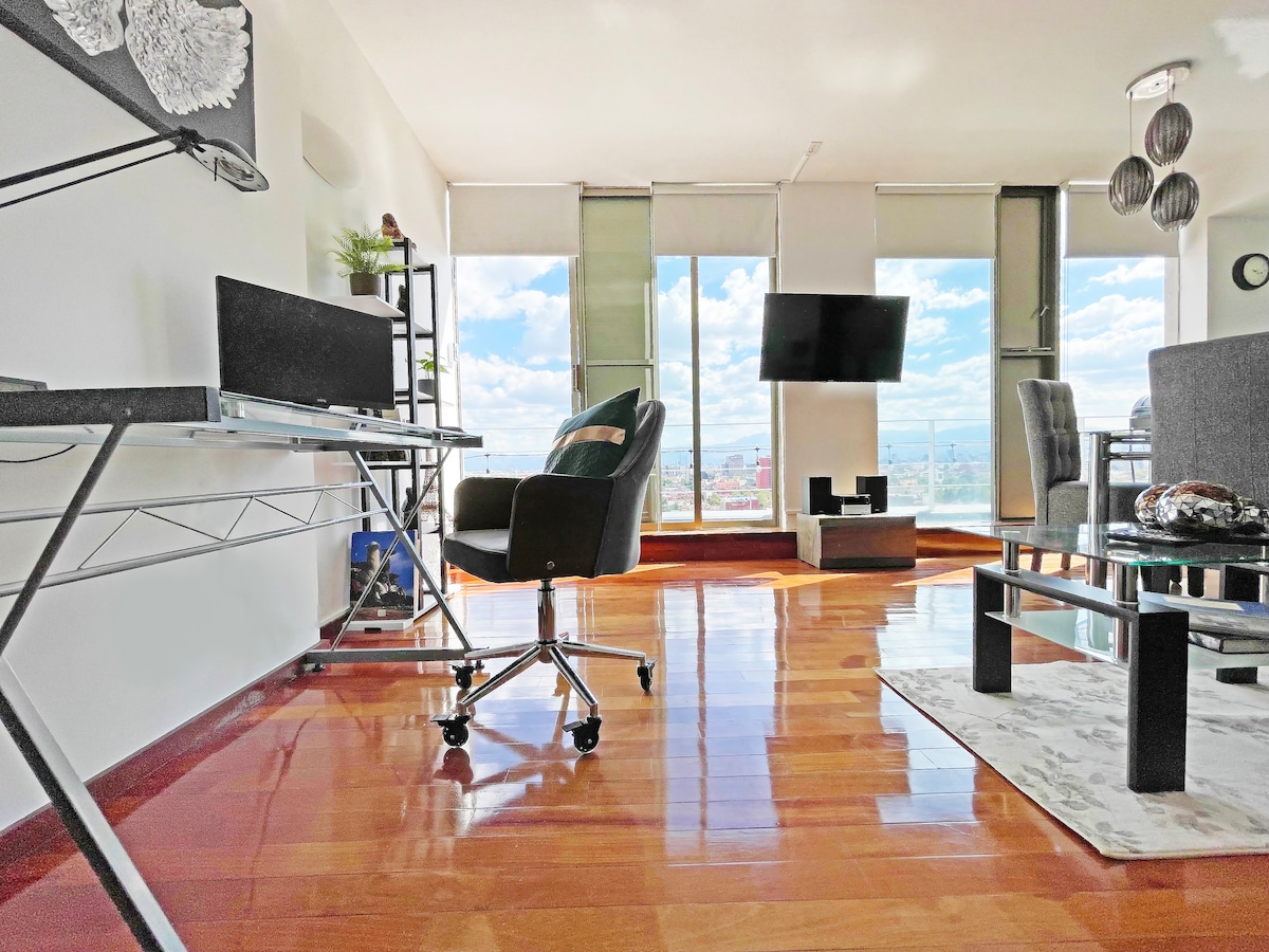 A modern workspace is presented with a sleek desk and ergonomic chair facing floor-to-ceiling windows. A 50-inch TV is mounted on the wall, and light reflects off the polished wooden floor. Nearby, a cozy seating area includes a glass coffee table and two chairs.