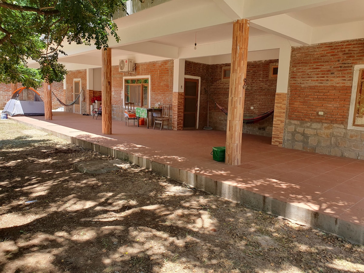 A spacious covered patio is supported by wooden columns, featuring areas for seating and relaxation. The terracotta tile flooring extends under a canopy of trees, providing shade. A hammock is visible, and an outdoor kitchen setup is indicated by the nearby furniture.