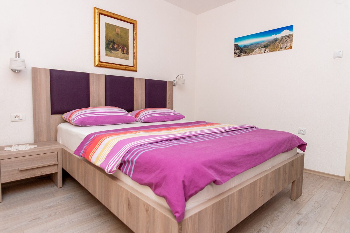 A cozy bedroom features a spacious bed adorned with vibrant purple and striped bedding. Simple bedside lamps are mounted on the wall, and a wooden nightstand holds decorative elements. A framed landscape artwork adds a touch of character to the neutral-colored walls.