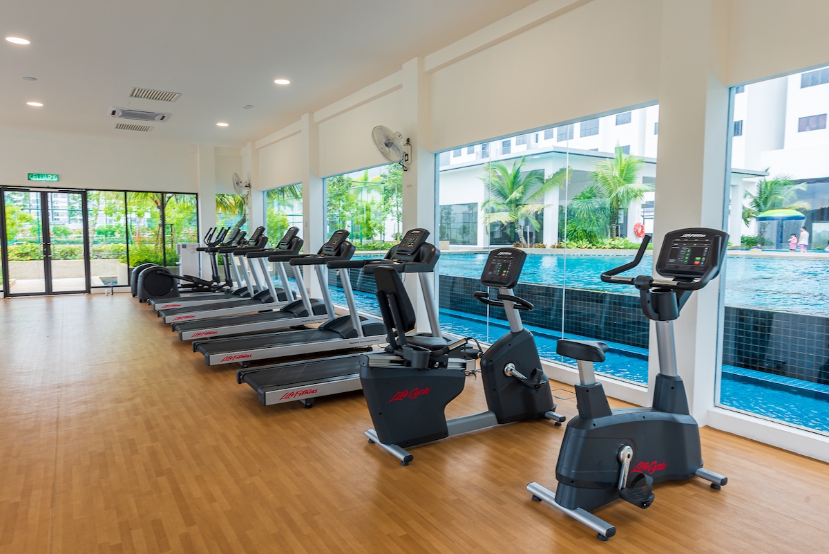 A well-equipped gym features multiple treadmills and stationary bikes, all facing large windows that overlook a tranquil outdoor swimming pool. The space is illuminated by natural light, enhancing the workout environment.