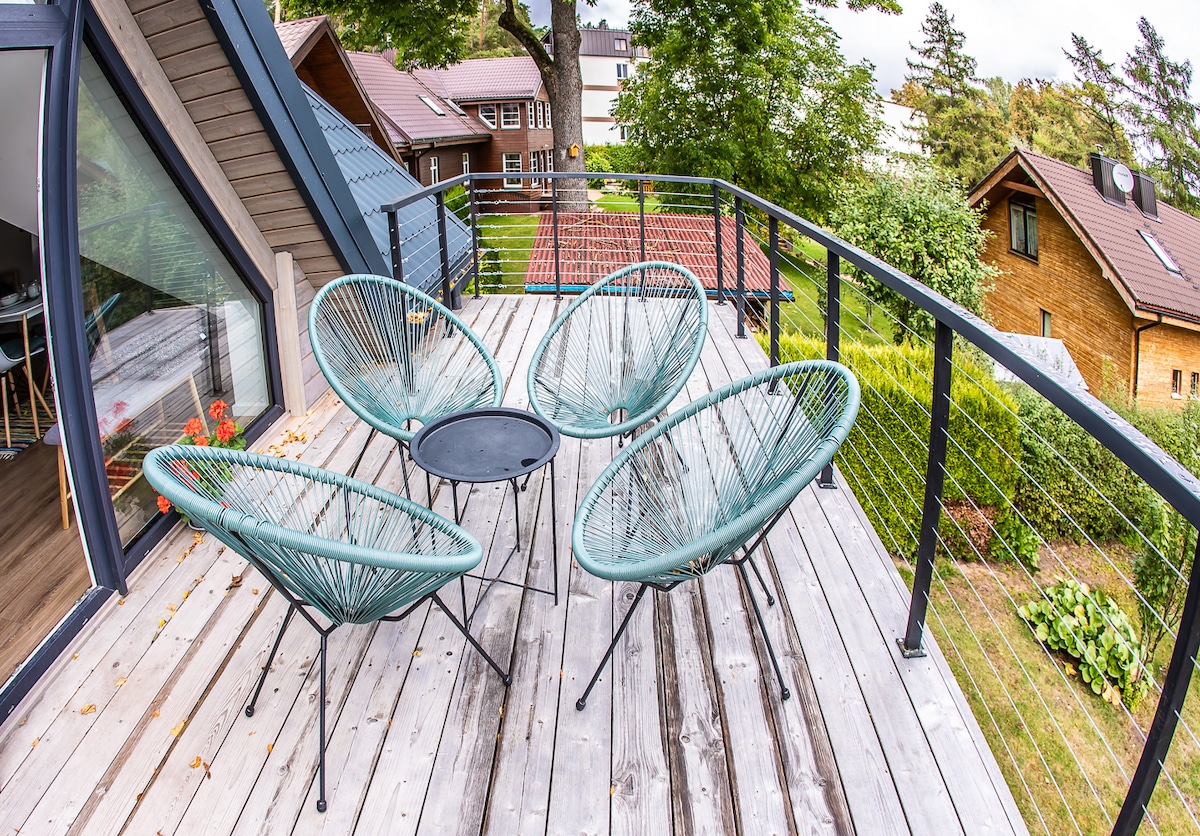 A spacious terrace is equipped with four round, green chairs arranged around a small black table. The wooden deck is framed by greenery and offers a view of surrounding homes and trees, providing a tranquil outdoor space.